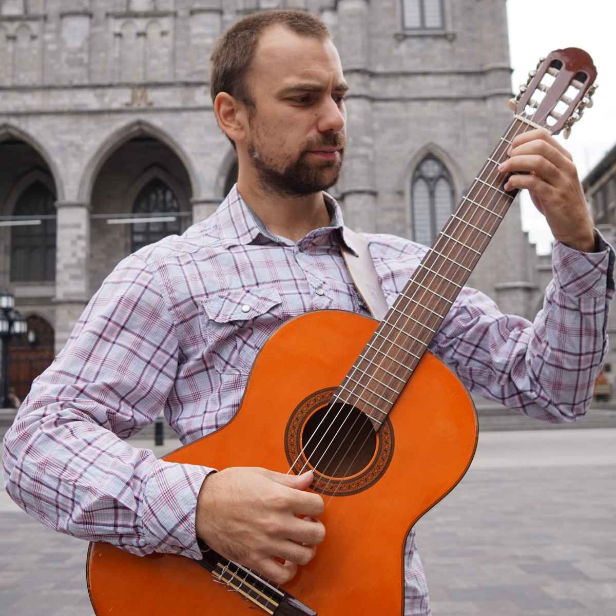 Brett wearing a plaid shirt playing an orange classical guitar at the Place D'armes in Old Montreal